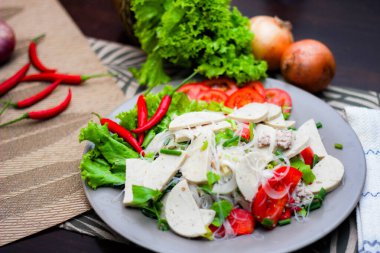 Gri bir tabakta servis edilen Vietnam sosisli Baharatlı Cam Erişte Salatası Ahşap masa ve çeşitli sebzeler.