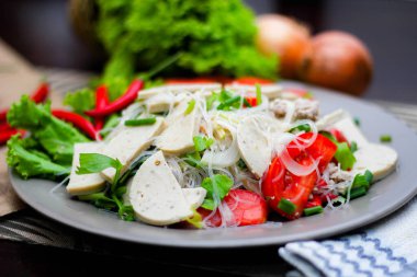 Gri bir tabakta servis edilen Vietnam sosisli Baharatlı Cam Erişte Salatası Ahşap masa ve çeşitli sebzeler.