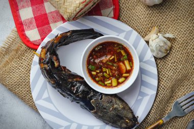 Izgara yayın balığının yer aldığı görsel olarak çekici bir kompozisyon. Balık altın kahverengidir ve daldırma sosunun canlı kırmızısıyla çok güzel tezat oluşturur. Kırsal ahşap çatal bıçak takımı ve dokuma örtüsü görüntüye biraz özgünlük katıyor..