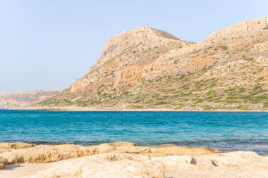 Bu manzara fotoğrafı Avrupa 'da, Yunanistan' da, Girit 'te, Balos' ta, Akdeniz 'in kıyısında, yazın çekildi. Kayalık kayalıkların dibinde, güneşin altında pembe yansımaları olan kumlu bir sahil görüyoruz..