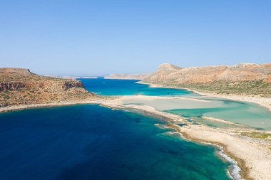 Bu manzara fotoğrafı Avrupa 'da, Yunanistan' da, Girit 'te, Balos' ta, Akdeniz 'in kıyısında, yazın çekildi. Kayalık kayalıkların dibinde, güneşin altında pembe yansımaları olan kumlu bir sahil görüyoruz..