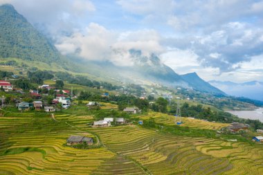 Bu manzara fotoğrafı Asya 'da, Vietnam' da, Tonkin 'de, Sapa' da Lao Cai 'de, yazın çekildi. Yeşil ve sarı pirinç tarlalarıyla geleneksel köyü görüyoruz. Yeşil dağlarda, bulutların altında..