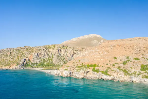 This landscape photo was taken, in Europe, in Greece, in Crete, in Preveli, By the Mediterranean sea, in summer. We see the sandy beach at the foot of the mountain next to a green palm grove, under the sun.