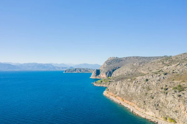 This landscape photo was taken, in Europe, in Greece, in the Peloponnese, in Argolis, in Nafplio, at the seaside of Myrto, in summer. We see the arid rocky coast and its green countryside at the edge of small beaches, under the sun.