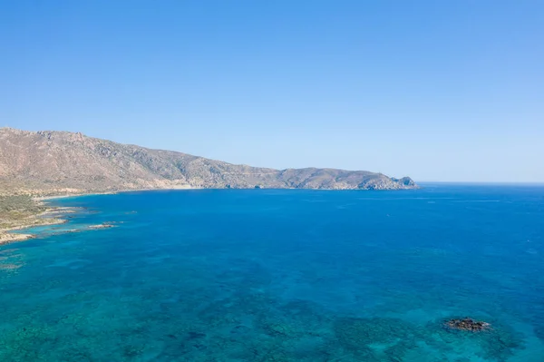 This landscape photo was taken, in Europe, in Greece, in Crete, in Elafonisi, By the Mediterranean sea, in summer. We see the rocky coast and its arid mountains, under the sun.