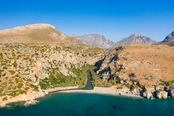 Это пейзажное фото было сделано в Европе, Греции, на Крите, в Превели, у Средиземного моря, летом. Мы видим песчаный пляж у подножия горы рядом с зеленой пальмовой рощей, под солнцем.