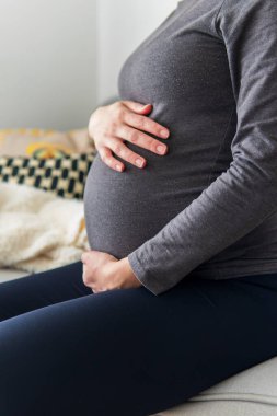 Hamile kadın koltukta rahatça oturup ellerini karnının üstüne koyup dinleniyor. Hamilelik acısı ve yorgunluğu yüzünden dinlenen bir kadın. Annelik, hamilelik kavramı. Ön çekim..
