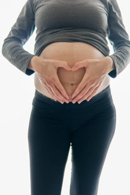 Hamile bir kadın pencerenin önünde duruyor ve elleri karnının üzerinde kalp şeklinde duruyor. Pencereden gelen parlak ışık, kadını ve güzel hamileliğini yansıtıyor..