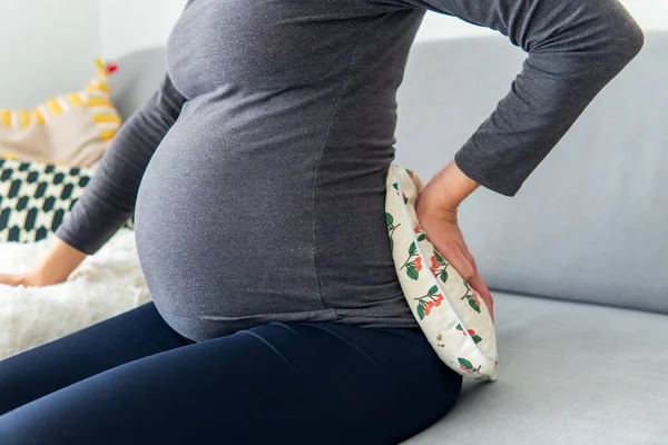 孕妇舒舒服服地坐在沙发上 下背上抱着一个治疗用的热枕头 同时抱着她的肚子 怀孕期间的背痛治疗和使用暖气垫缓解疼痛 — 图库照片