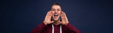 Kırmızı kazaklı, mavi arka planda bir adam elleri yüzünün yanında bağırıyor.