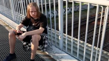 Genç bir adam tren istasyonunda bir köprüde oturuyor.