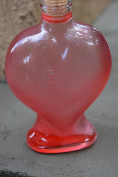A beautiful glass vessel in the shape of a heart with a light wooden stopper and a red astringent liquid stands on the street.