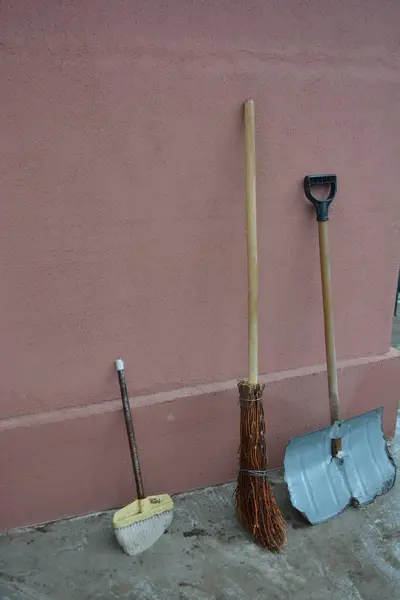 Equipment, household items for housework. An old worn-out broom, a worn-out metal shovel, a sweeping brush standing under the house.
