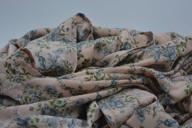 Alışılmadık, parlak yaz kumaşı. Beyaz, ince sentetik, mavi çiçekli ipek kumaş, yeşil yapraklar beyaz plastik arka planda yer almaktadır..