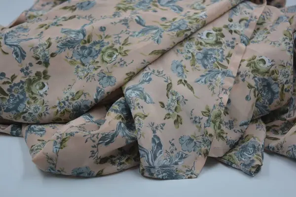 Alışılmadık, parlak yaz kumaşı. Beyaz, ince sentetik, mavi çiçekli ipek kumaş, yeşil yapraklar beyaz plastik arka planda yer almaktadır..