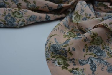 Alışılmadık, parlak yaz kumaşı. Beyaz, ince sentetik, mavi çiçekli ipek kumaş, yeşil yapraklar beyaz plastik arka planda yer almaktadır..