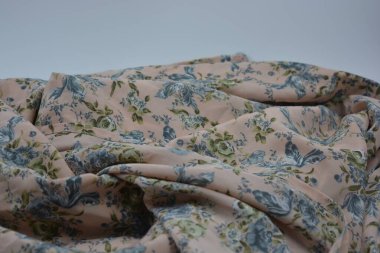 Alışılmadık, parlak yaz kumaşı. Beyaz, ince sentetik, mavi çiçekli ipek kumaş, yeşil yapraklar beyaz plastik arka planda yer almaktadır..