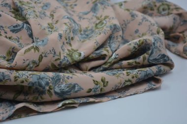 Alışılmadık, parlak yaz kumaşı. Beyaz, ince sentetik, mavi çiçekli ipek kumaş, yeşil yapraklar beyaz plastik arka planda yer almaktadır..