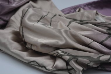 Alışılmadık, parlak ve parıltılı yaz kumaşı. Bej, kum, narin mor, ince sentetik, koyu renk göz kapakları olan ipek kumaş, beyaz plastik arka planda çizgiler var..