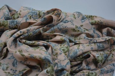 Alışılmadık, parlak yaz kumaşı. Beyaz, ince sentetik, mavi çiçekli ipek kumaş, yeşil yapraklar beyaz plastik arka planda yer almaktadır..