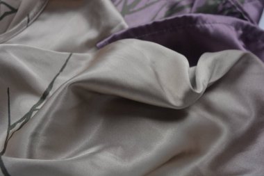 Alışılmadık, parlak ve parıltılı yaz kumaşı. Bej, kum, narin mor, ince sentetik, koyu renk göz kapakları olan ipek kumaş, beyaz plastik arka planda çizgiler var..