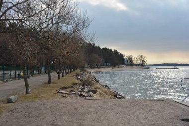 Finlandiya, Espoo 'da körfez ve bahar doğası olan parlak ve renkli manzaralar. Güzel berrak su, sarı kumlu büyük sahil, dalgalar, uzun yeşil çam ağaçları, bahçe ağaçları, büyük taşlar, renkli su deposu, körfez, göl.