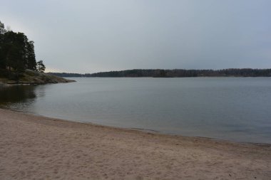 Finlandiya, Espoo 'da körfez ve bahar doğası olan parlak ve renkli manzaralar. Güzel berrak su, sarı kumlu büyük sahil, dalgalar, uzun yeşil çam ağaçları, bahçe ağaçları, büyük taşlar, renkli su deposu, körfez, göl.