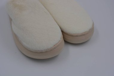 Güzel ve alışılmadık kadın eşyaları, günlük ev ayakkabıları. Kalın kahverengi, bej tabanlı sentetik kürkten yapılmış beyaz yumuşak terlikler beyaz plastik bir arka planda yer almaktadır..