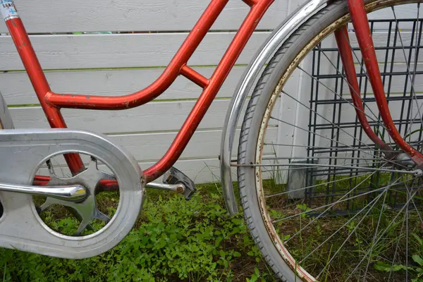 Eşyalar, eşyalar, spor, sağlık. Eski bir kırmızı metal bisiklet yeşil bir çimenlikte beyaz bir çite karşı duruyor. Bisiklet parçaları eski, bisiklet tekerlekleri paslı, bisikletin metal kısımlarında birçok kırmızı pas lekesi var..