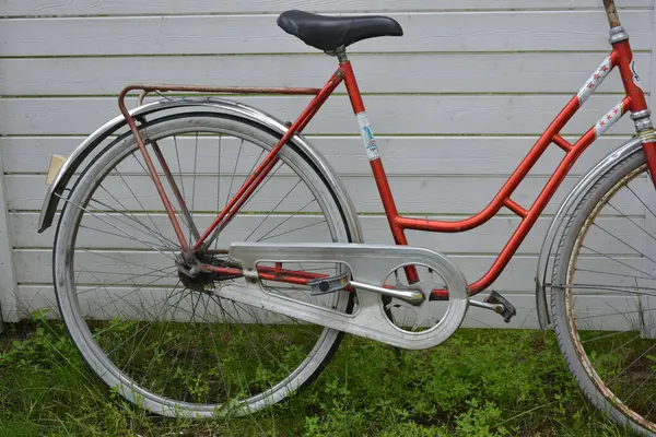 Eşyalar, eşyalar, spor, sağlık. Eski bir kırmızı metal bisiklet yeşil bir çimenlikte beyaz bir çite karşı duruyor. Bisiklet parçaları eski, bisiklet tekerlekleri paslı, bisikletin metal kısımlarında birçok kırmızı pas lekesi var..