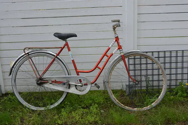 Eşyalar, eşyalar, spor, sağlık. Eski bir kırmızı metal bisiklet yeşil bir çimenlikte beyaz bir çite karşı duruyor. Bisiklet parçaları eski, bisiklet tekerlekleri paslı, bisikletin metal kısımlarında birçok kırmızı pas lekesi var..