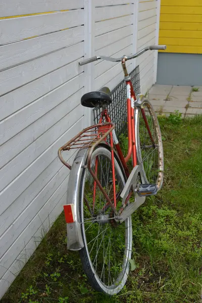 Eşyalar, eşyalar, spor, sağlık. Eski bir kırmızı metal bisiklet yeşil bir çimenlikte beyaz bir çite karşı duruyor. Bisiklet parçaları eski, bisiklet tekerlekleri paslı, bisikletin metal kısımlarında birçok kırmızı pas lekesi var..