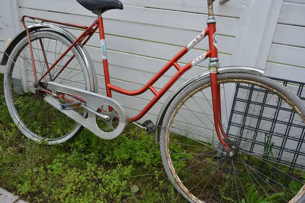 Eşyalar, eşyalar, spor, sağlık. Eski bir kırmızı metal bisiklet yeşil bir çimenlikte beyaz bir çite karşı duruyor. Bisiklet parçaları eski, bisiklet tekerlekleri paslı, bisikletin metal kısımlarında birçok kırmızı pas lekesi var..