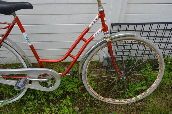 Eşyalar, eşyalar, spor, sağlık. Eski bir kırmızı metal bisiklet yeşil bir çimenlikte beyaz bir çite karşı duruyor. Bisiklet parçaları eski, bisiklet tekerlekleri paslı, bisikletin metal kısımlarında birçok kırmızı pas lekesi var..