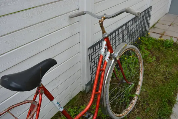 Eşyalar, eşyalar, spor, sağlık. Eski bir kırmızı metal bisiklet yeşil bir çimenlikte beyaz bir çite karşı duruyor. Bisiklet parçaları eski, bisiklet tekerlekleri paslı, bisikletin metal kısımlarında birçok kırmızı pas lekesi var..