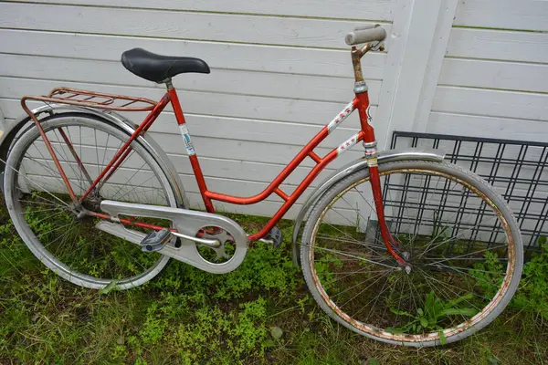 Eşyalar, eşyalar, spor, sağlık. Eski bir kırmızı metal bisiklet yeşil bir çimenlikte beyaz bir çite karşı duruyor. Bisiklet parçaları eski, bisiklet tekerlekleri paslı, bisikletin metal kısımlarında birçok kırmızı pas lekesi var..