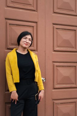 Güzel Asyalı kadın (LGBTQ) doğal makyaj, moda sarı deri kıyafetler ve gözlüklerle eski kentte ve klasik bina modasında poz verir.