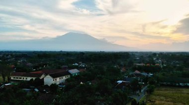 Gün batımında bulutlu bir gökyüzünün altında arka planda bir dağ olan kırsal bir arazinin havadan görünüşü.
