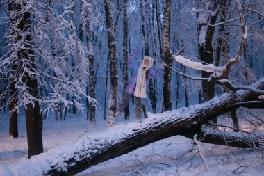 Küçük kız karlı bir ormanda devrilmiş bir ağaçta duruyor.