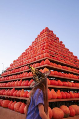 10 years girl in witch hat with pumpkin pyramid in background clipart