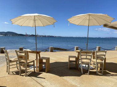 Sıcak yaz mevsiminde okyanusun önünde yemek bekleyen sandalyeli ve şemsiyeli iki eski ahşap masa.