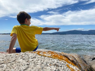 Sarı tişörtlü genç deniz kenarındaki bir kayada oturuyor ve parmağıyla işaret ediyor.