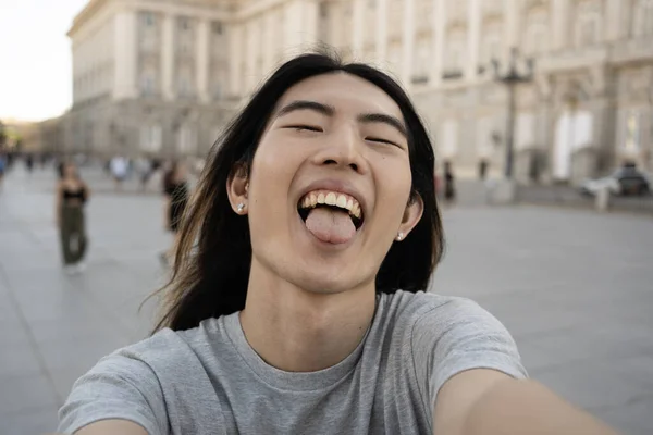 Asian young guy showing his tongue while taking selfie photo in Madrid city.