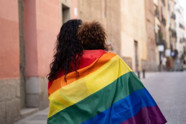 Şehir sokağında gökkuşağı bayrağıyla kaplanmış iki kadının arka görüntüsü. Lgbt aşk gurur konsepti.