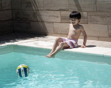 Dominikli genç çocuk tatillerde havuz başında oturup top oyuncağına bakıyor..