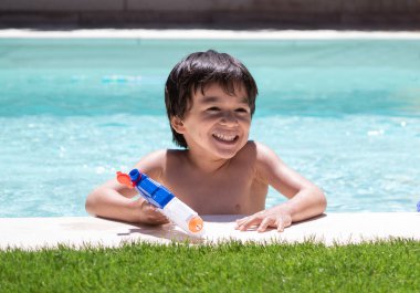 Havuzda otururken oyuncak silah tutan gülümseyen genç bir çocuk. Çocuklar için yaz planı kavramı.