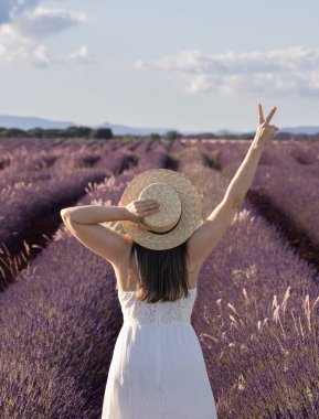 Hasır şapkalı bir kadın lavanta tarlasında duruyor. Ellerini havaya kaldırıyor ve barış işareti yapıyor.