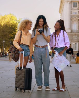 Üç genç kadın kaldırımda dikiliyorlar, biri elinde kamera. Günlük kıyafetler giyiyorlar ve bavul taşıyorlar. Sahne neşeli ve neşeli.