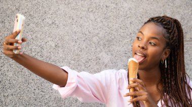 Gri arka planda izole edilmiş dondurma külahını yerken çekilmiş Afrikalı Amerikalı bir kadının fotoğrafı..