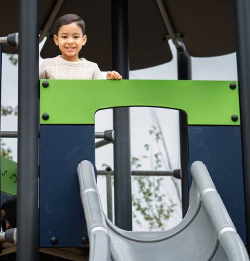 Çocuk oyun parkında kaydırağın tepesinde duruyor, aşağıya inmeden önce gülümsüyor.